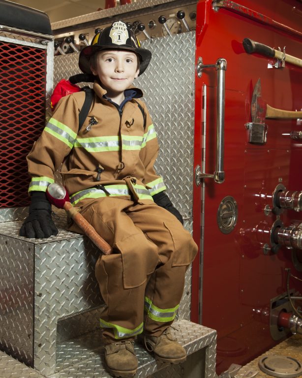 Young boy dressed as a fire fighter and sitting on a fire truck.